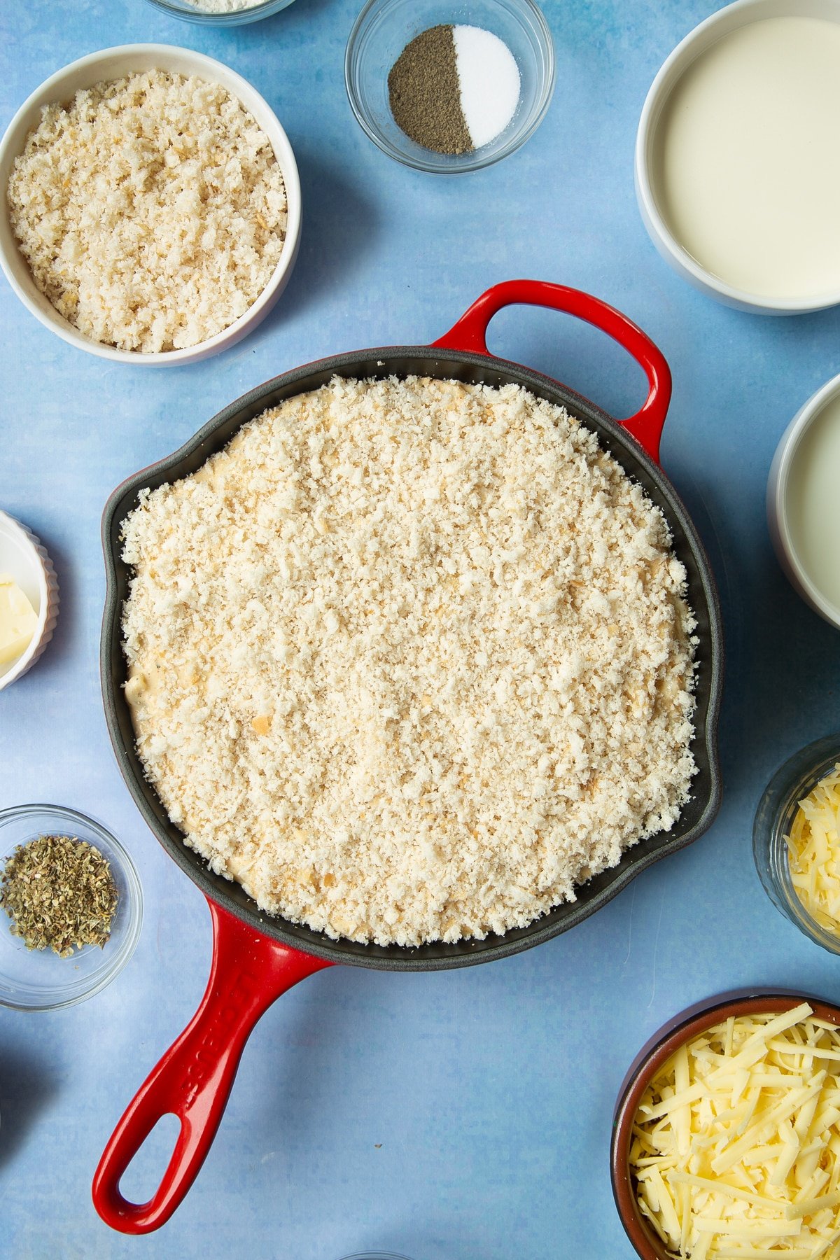 Spicy, cheesy, herby mac and cheese in a skillet with breadcrumbs on top. Ingredients to make garlic and herb mac and cheese surround the skillet.