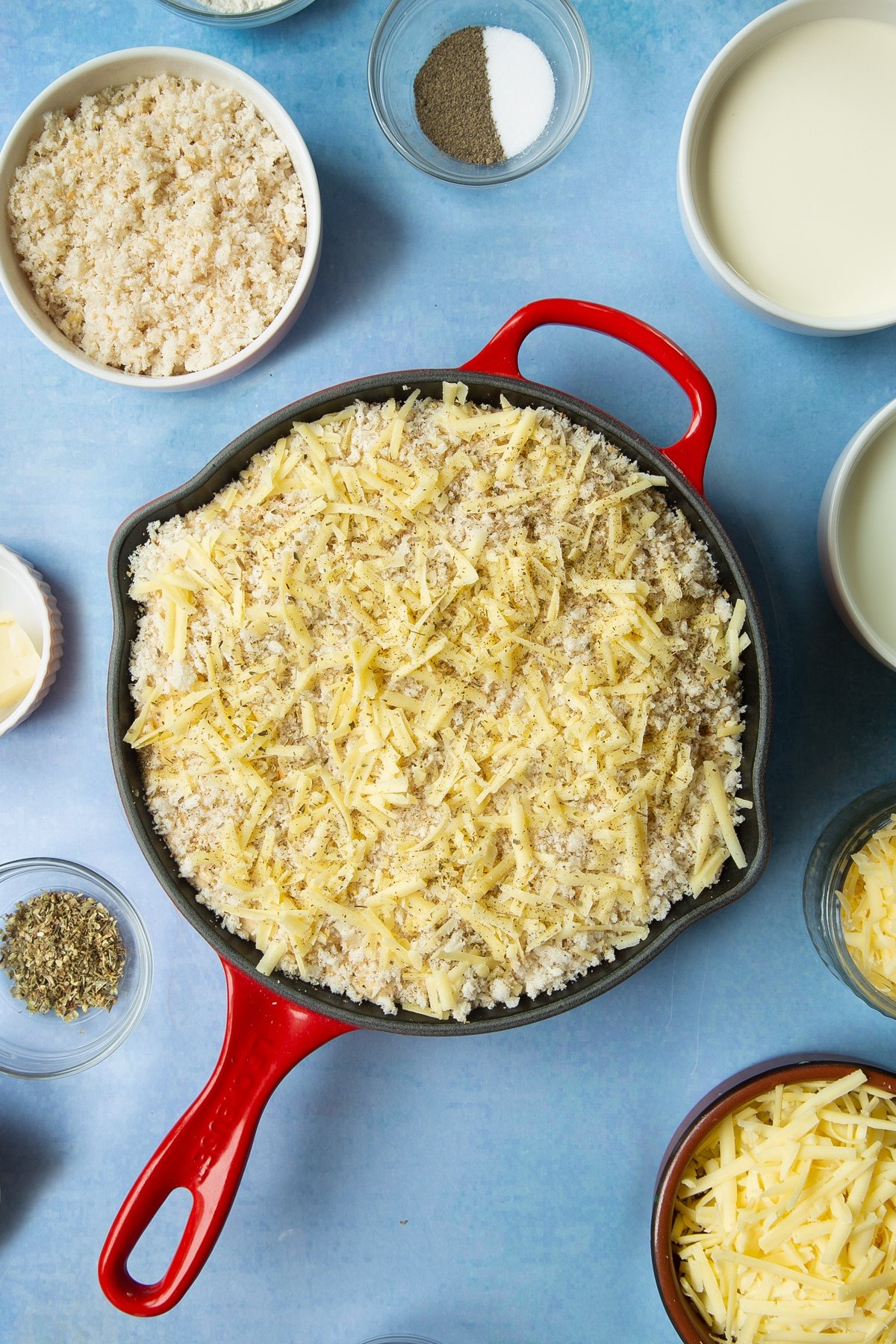 Spicy, cheesy, herby mac and cheese in a skillet with breadcrumbs, cheese and herbs on top. Ingredients to make garlic and herb mac and cheese surround the skillet.