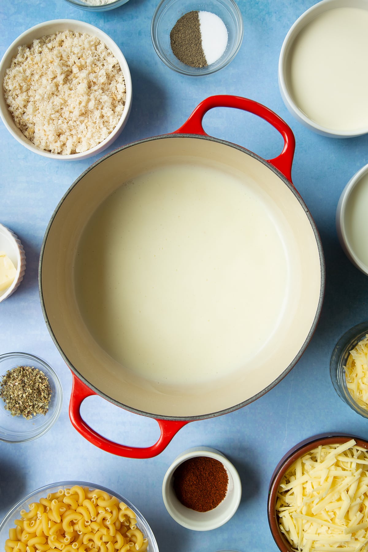 White sauce in a pan. Ingredients to make garlic and herb mac and cheese surround the pan.