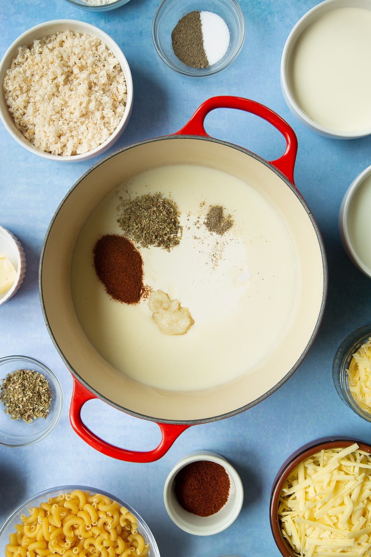 White sauce in a pan with paprika, oregano, garlic, salt and pepper on top. Ingredients to make garlic and herb mac and cheese surround the pan.