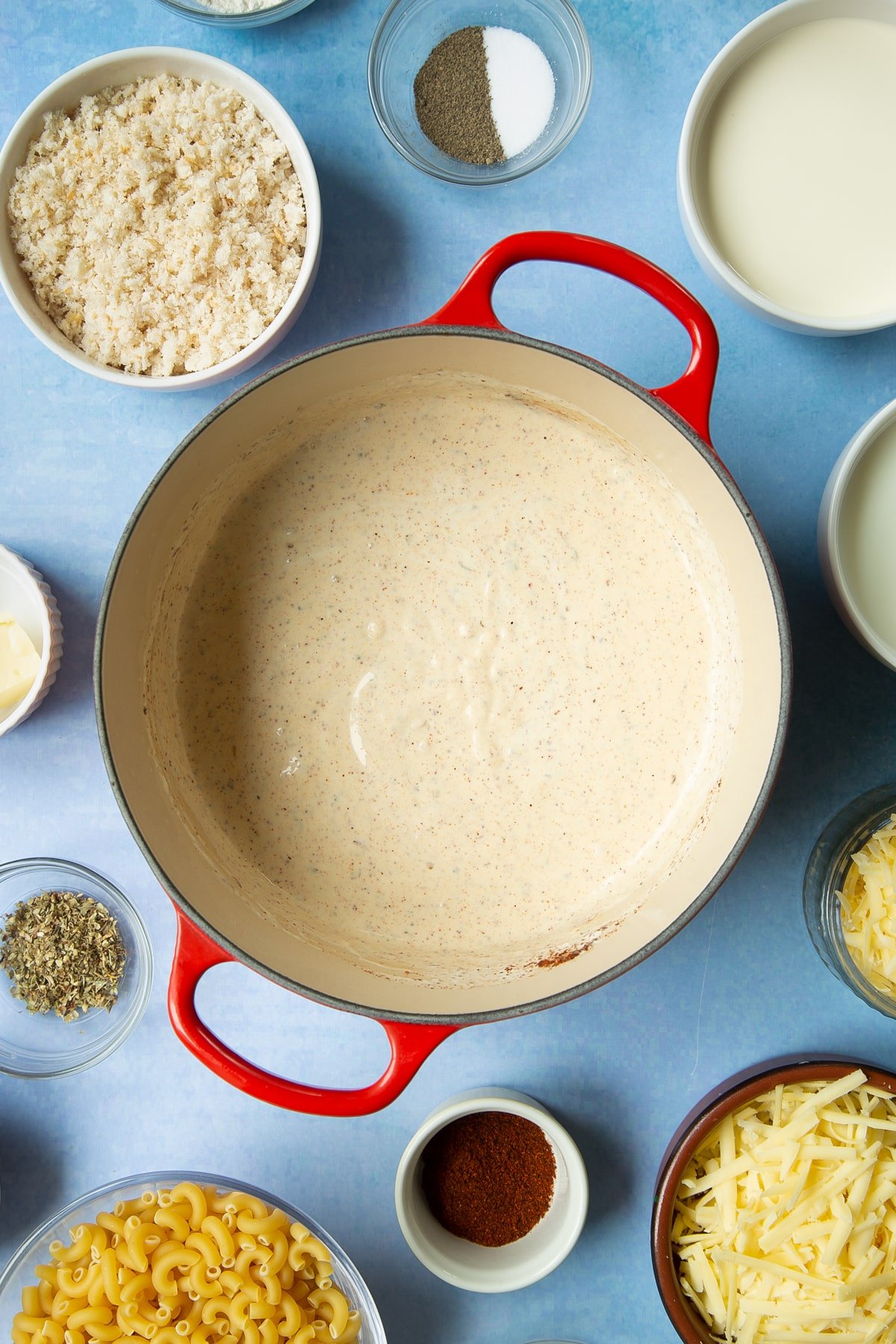 White sauce with paprika and herbs in a pan. Ingredients to make garlic and herb mac and cheese surround the pan.