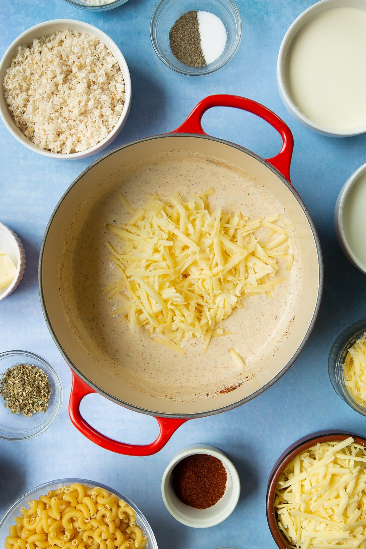 Spicy white sauce with grated cheese on top in a pan. Ingredients to make garlic and herb mac and cheese surround the pan.