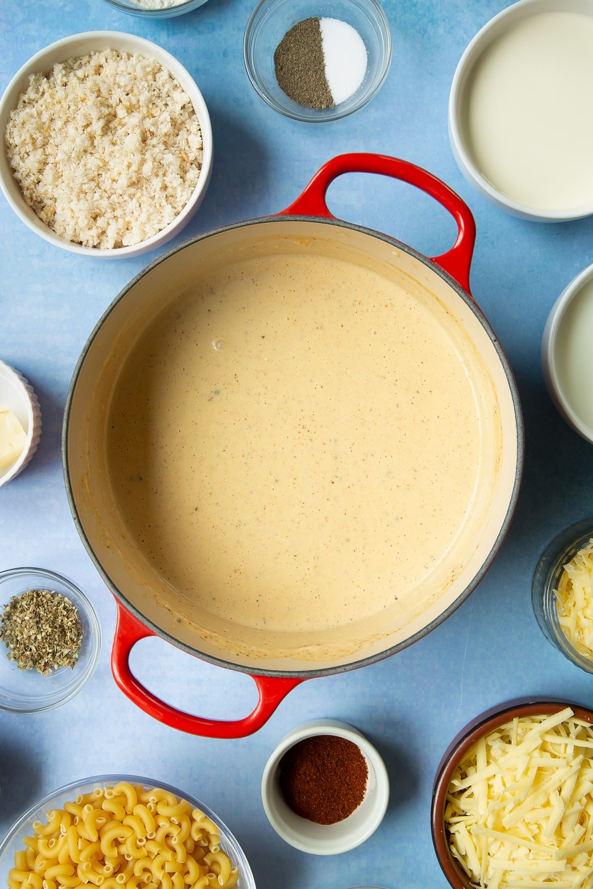 Spicy, cheesy white sauce in a pan. Ingredients to make garlic and herb mac and cheese surround the pan.