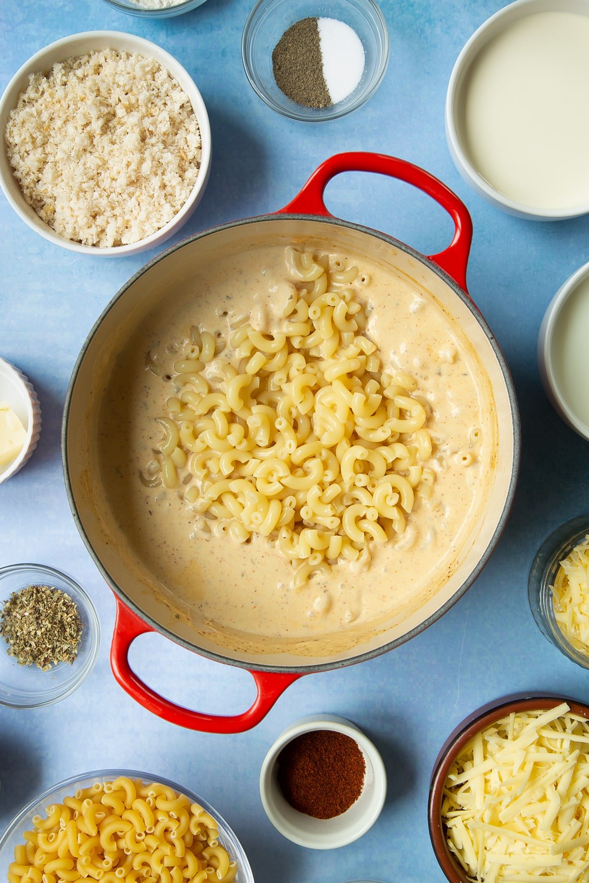 Spicy, cheesy white sauce with cooked macaroni on top in a pan. Ingredients to make garlic and herb mac and cheese surround the pan.