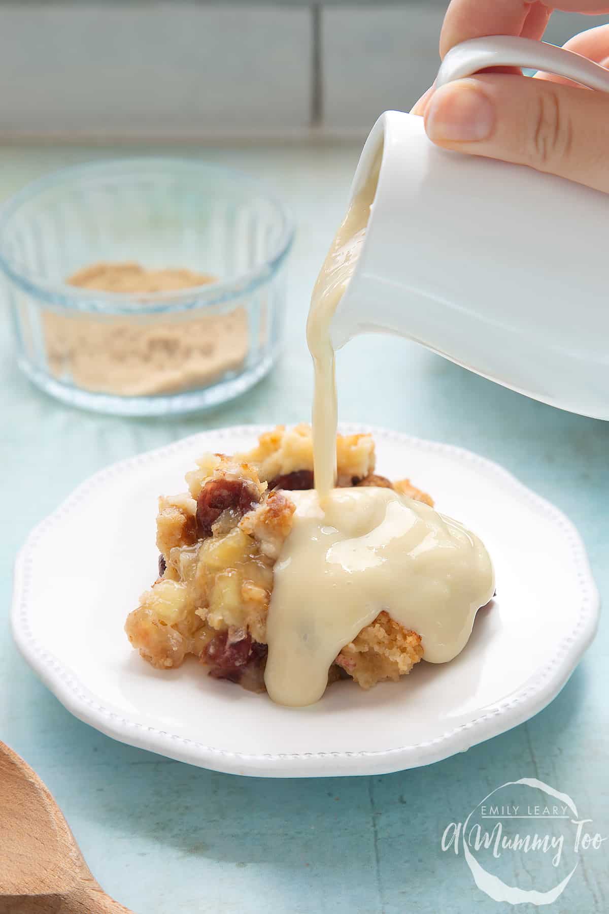 A berry crumble on a white plate covered with vegan custard being poured from a white jug.