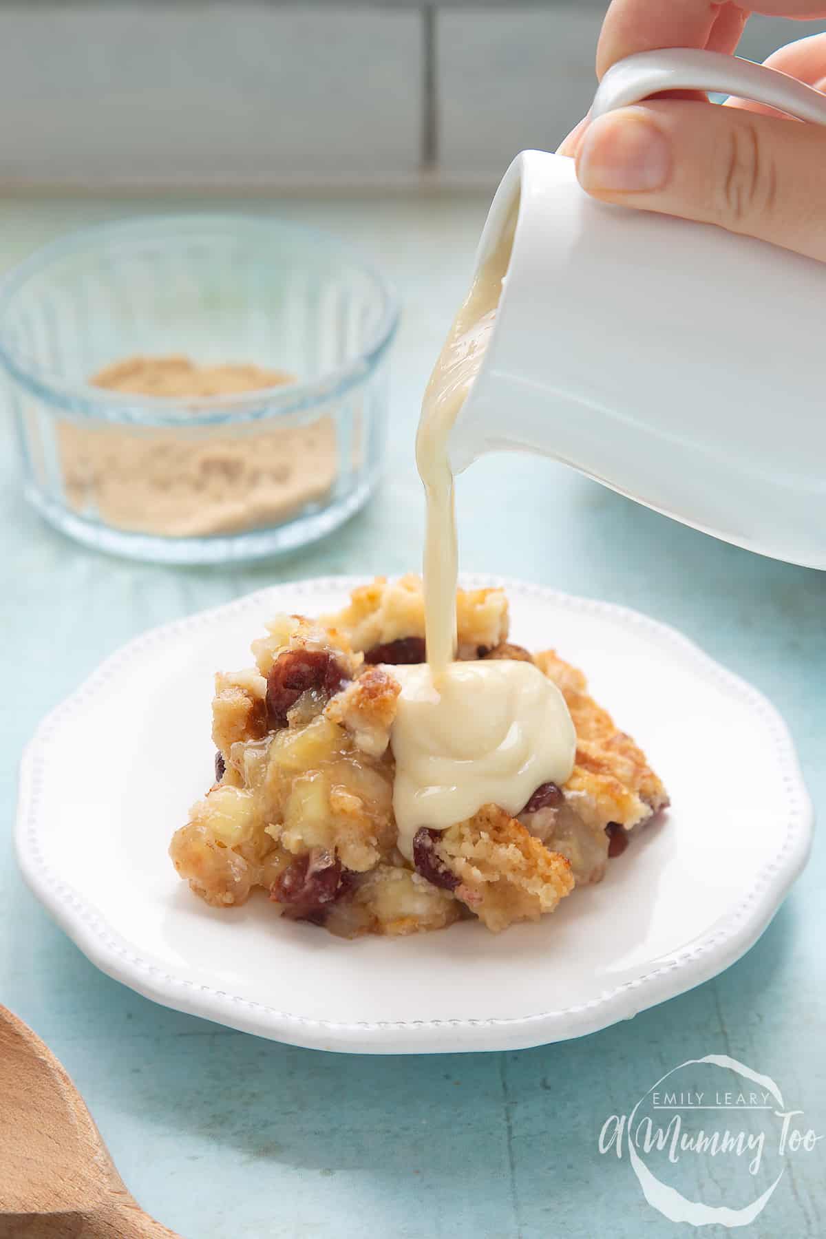 A berry crumble on a white plate. A hand holds a small white jug of vegan custard, which is being poured onto the crumble.