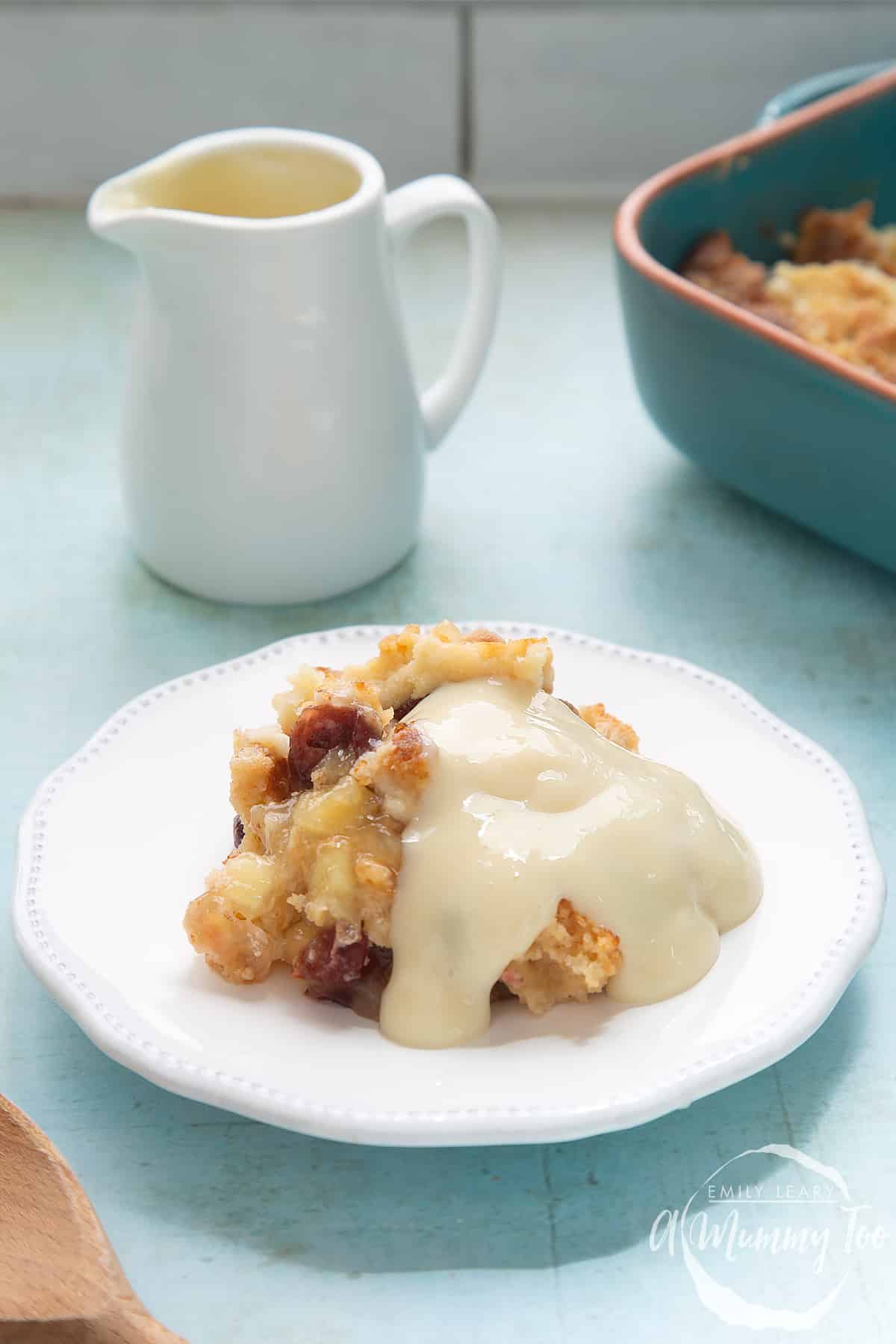 A berry crumble covered with vegan custard on a white plate. A small jug of vegan custard and a tray of crumble is shown in the background