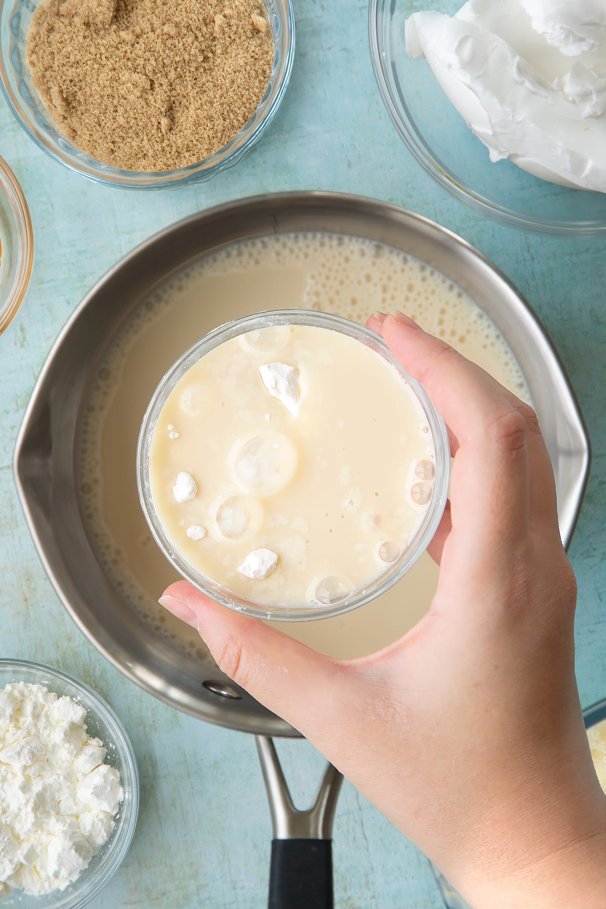 Soya milk, vanilla and sugar in a pan. Above it, a hand holds a small bowl filled with cornflour with milk. Ingredients to make vegan custard surround the pan.