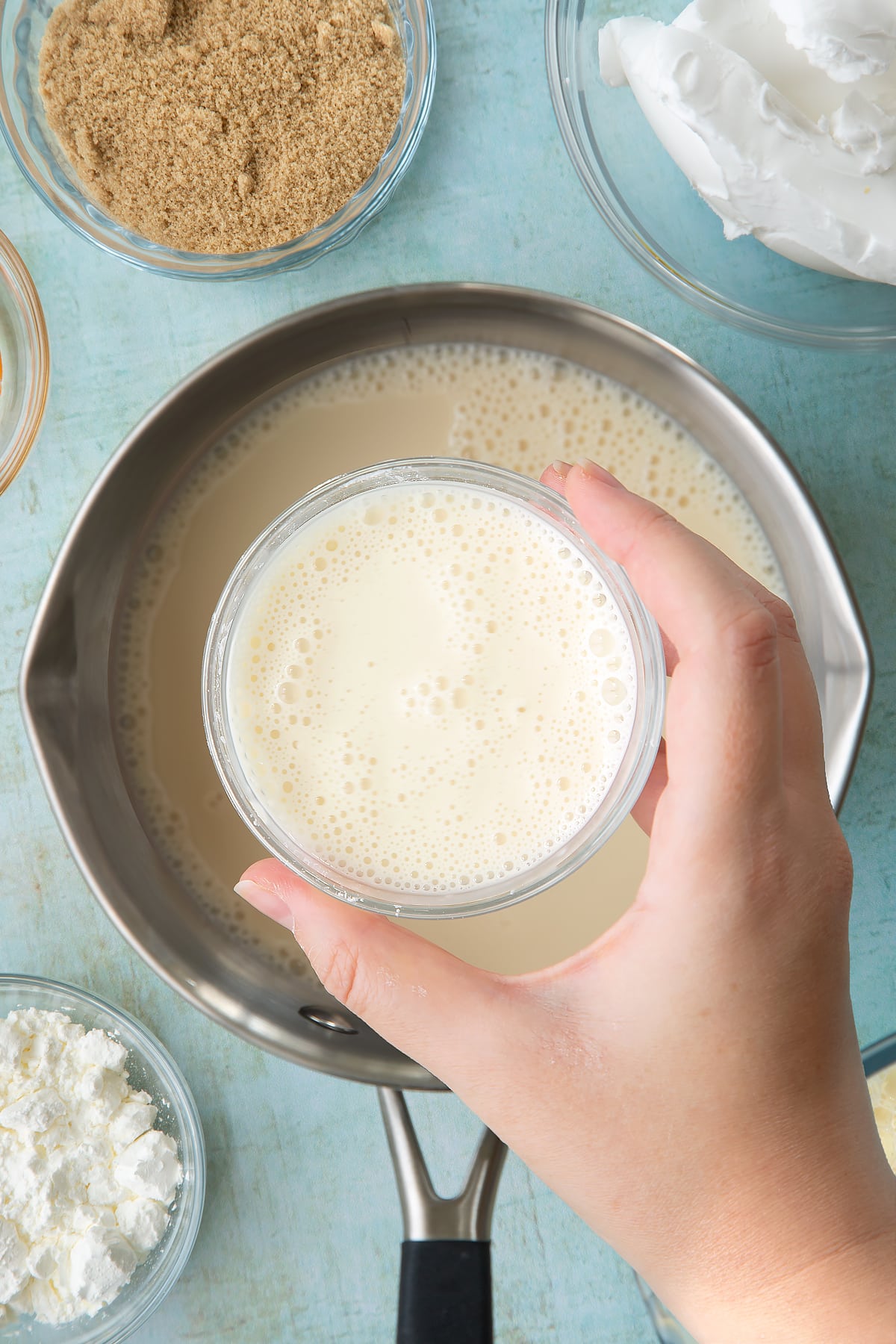 Soya milk, vanilla and sugar in a pan. Above it, a hand holds a small bowl filled with a mixture of cornflour and milk. Ingredients to make vegan custard surround the pan.