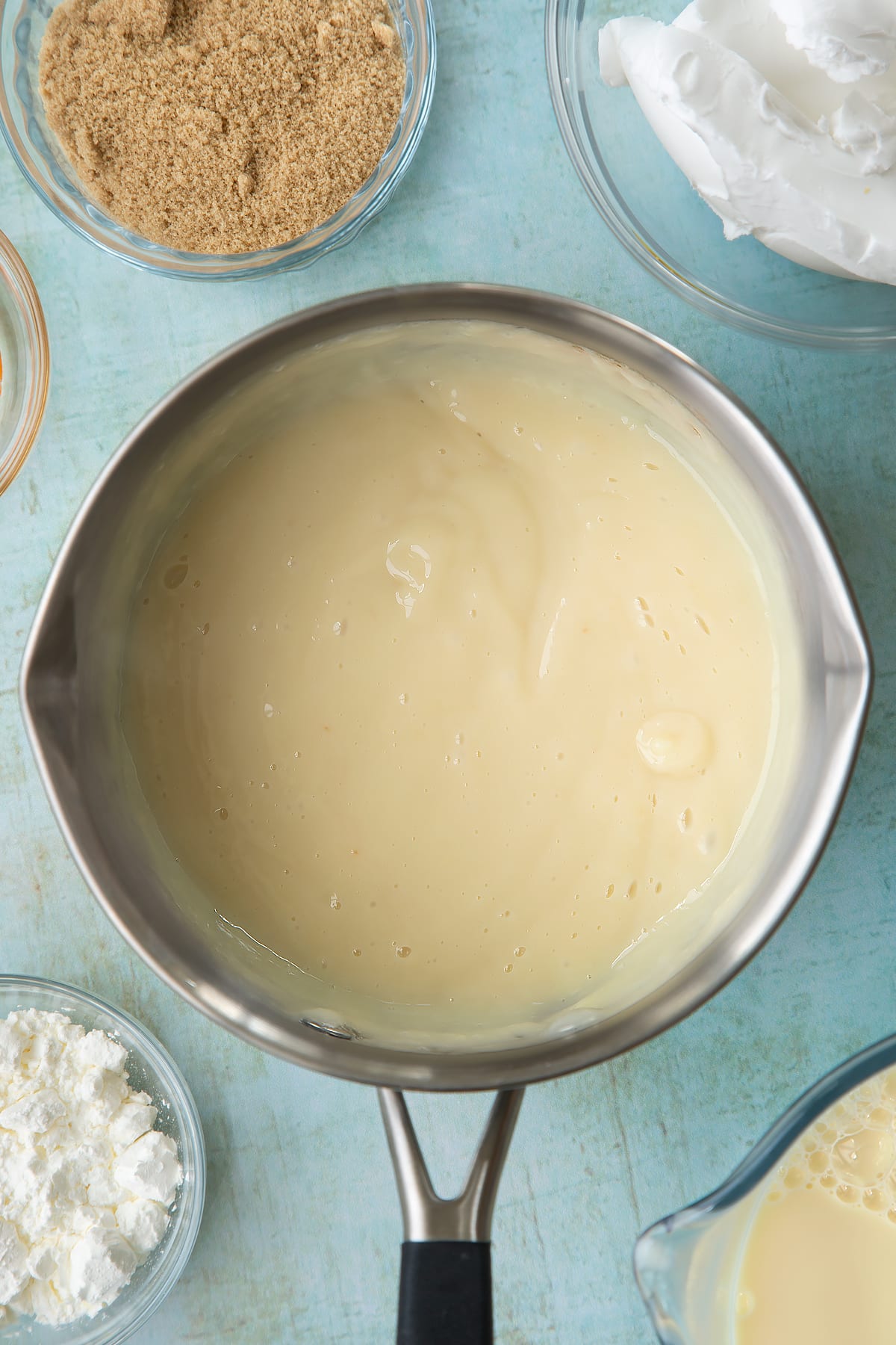 Soya milk, vanilla, sugar and cornflour thickened together in a pan. Ingredients to make vegan custard surround the pan.