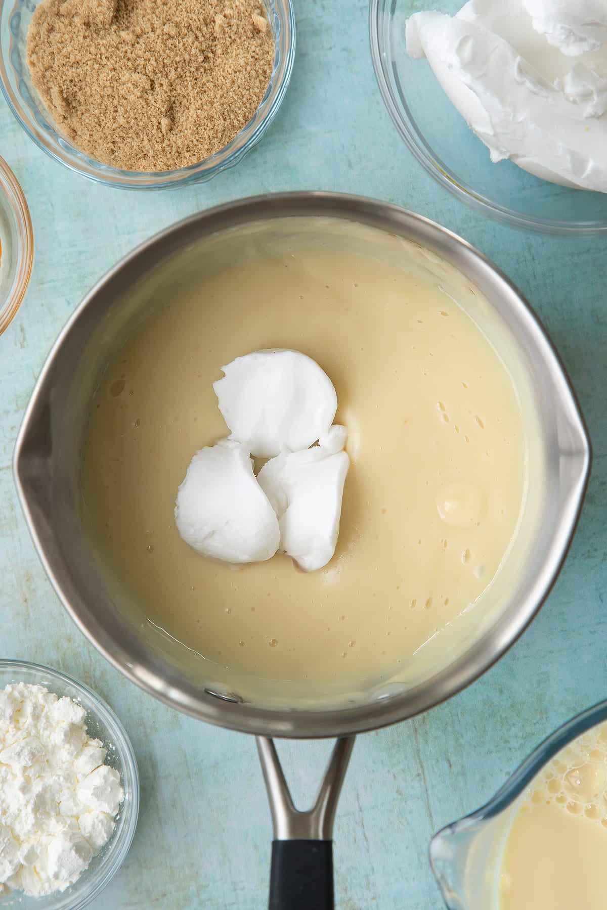 Soya milk, vanilla, sugar and cornflour thickened together in a pan with vegan cream cheese on top. Ingredients to make vegan custard surround the pan.