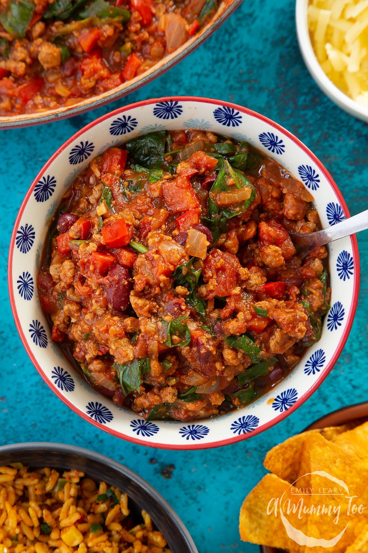 Vegetarian mince chilli in a bowl. A fork is in the bowl. Bowls containing tortilla chips, grated cheese and spicy rice are also shown.