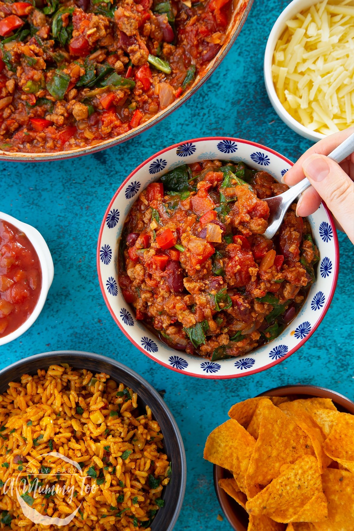 Vegetarian mince chilli in a bowl. A hand holding a fork delves into the chilli. Bowls containing tortilla chips, salsa, grated cheese and spicy rice are also shown.