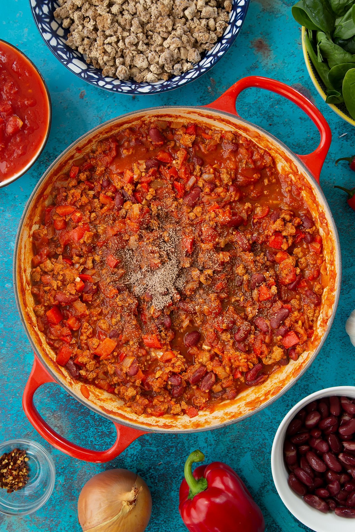 Vegetarian mince chilli in a pan with a pinch of salt and pepper on top. Ingredients to make vegetarian mince chilli surround the pan.