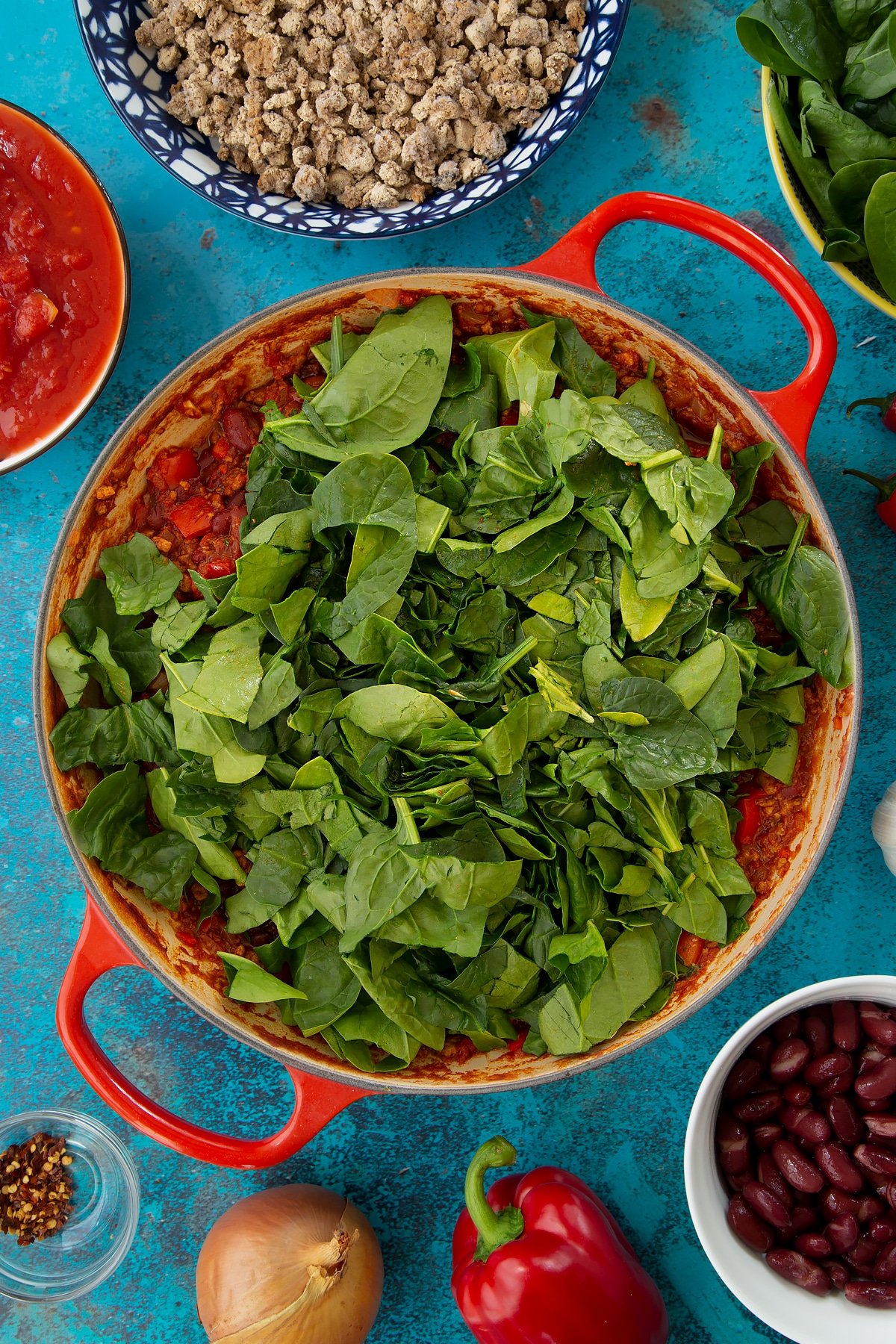 Vegetarian mince chilli in a pan with chopped fresh spinach on top. Ingredients to make vegetarian mince chilli surround the pan.