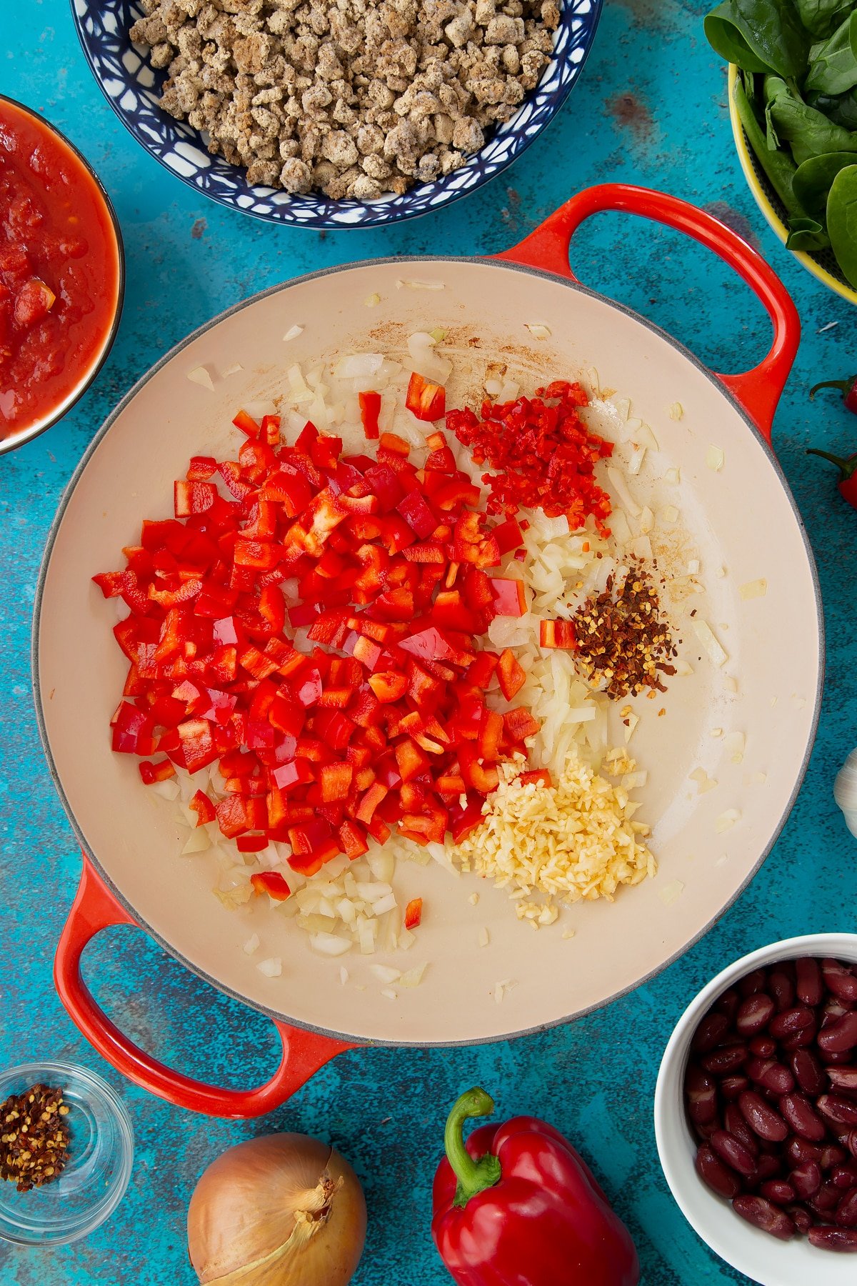 Lightly fried onion in a pan with minced garlic, chilli, chilli flakes and chopped red pepper. Ingredients to make vegetarian mince chilli surround the pan.