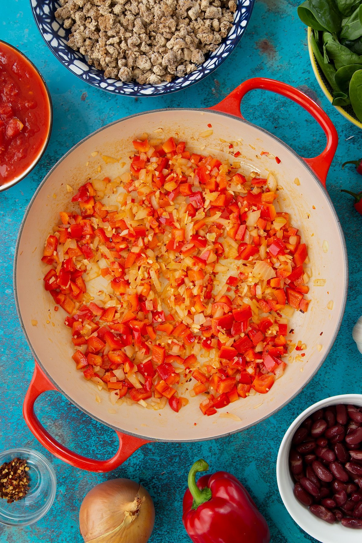 Lightly fried onion, minced garlic, chilli, chilli flakes and chopped red pepper in a pan. Ingredients to make vegetarian mince chilli surround the pan.