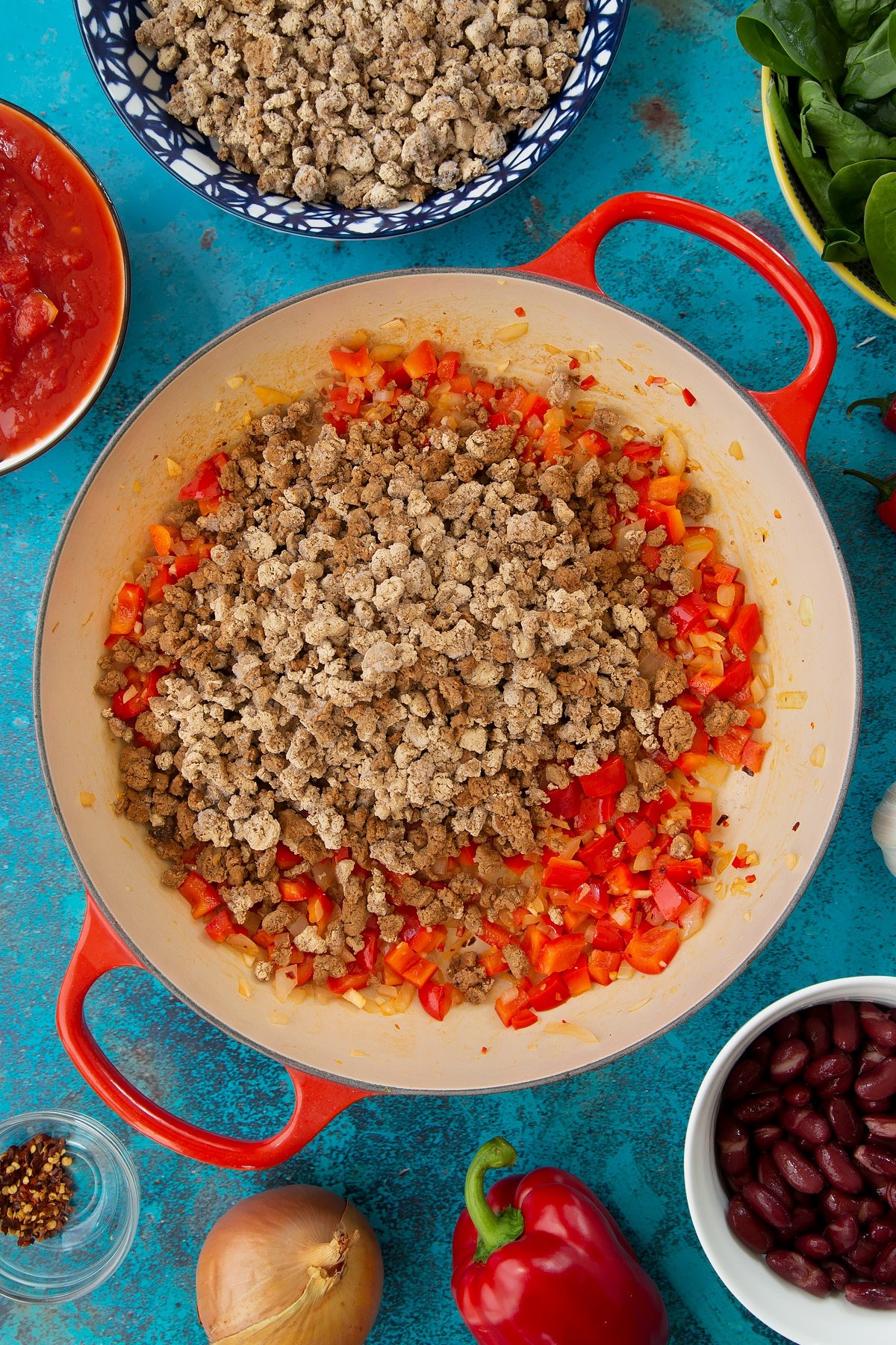 Lightly fried onion, minced garlic, chilli, chilli flakes and chopped red pepper in a pan with vegetarian mince on top. Ingredients to make vegetarian mince chilli surround the pan.