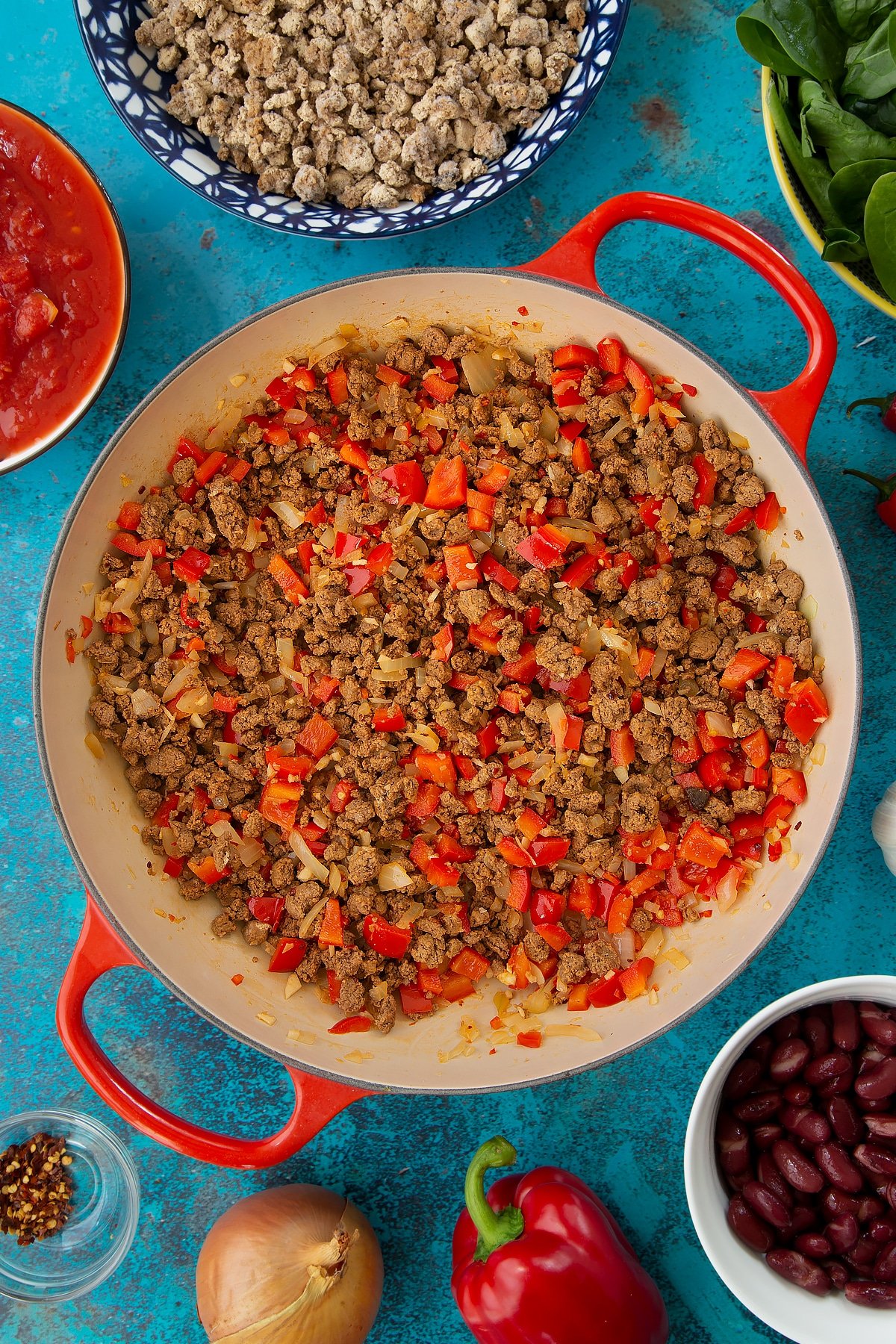 Fried onion, red pepper and vegetarian mince in a pan. Ingredients to make vegetarian mince chilli surround the pan.