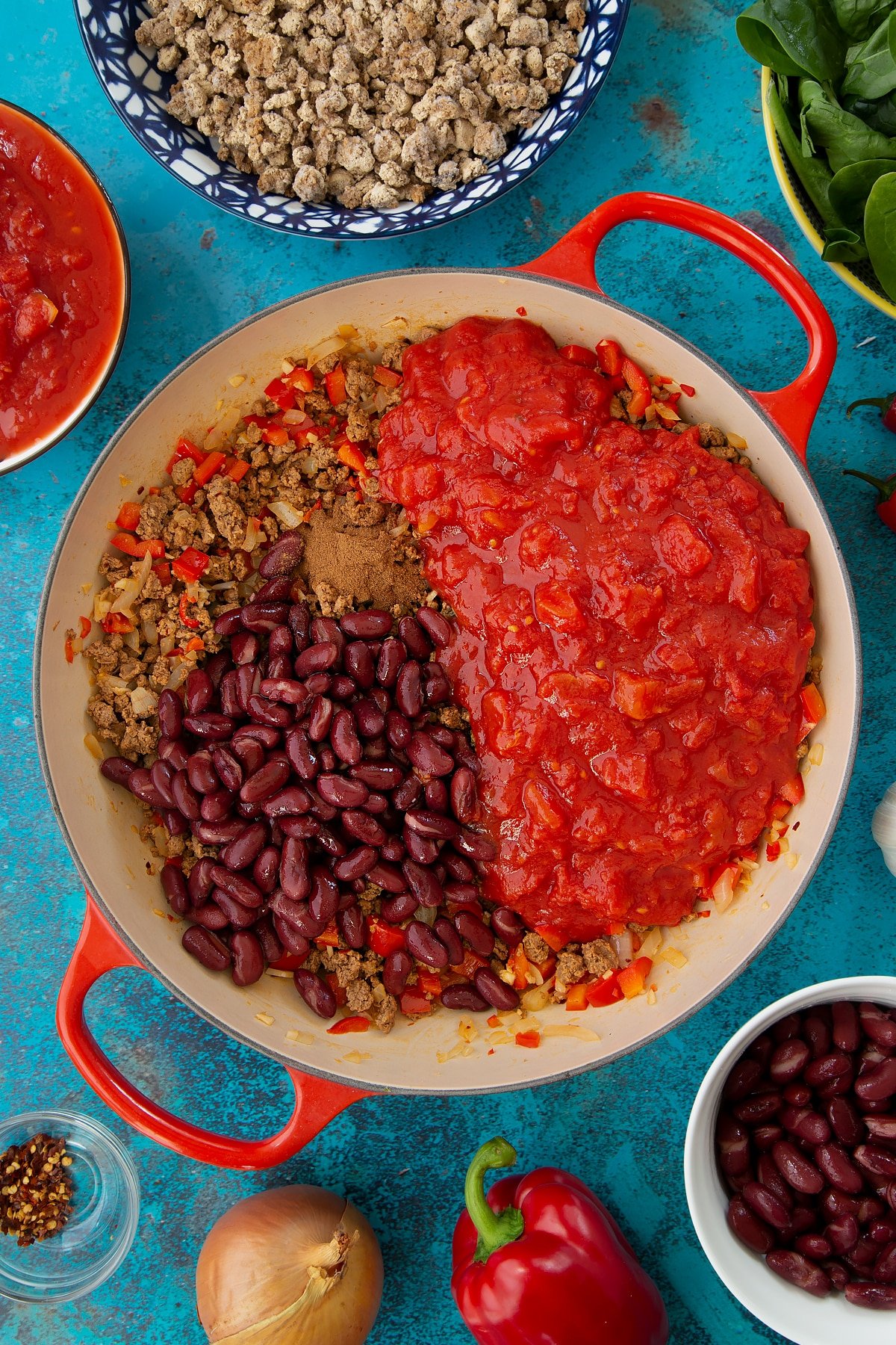 Fried onion, red pepper and vegetarian mince in a pan with kidney beans, tinned tomatoes and cinnamon. Ingredients to make vegetarian mince chilli surround the pan.