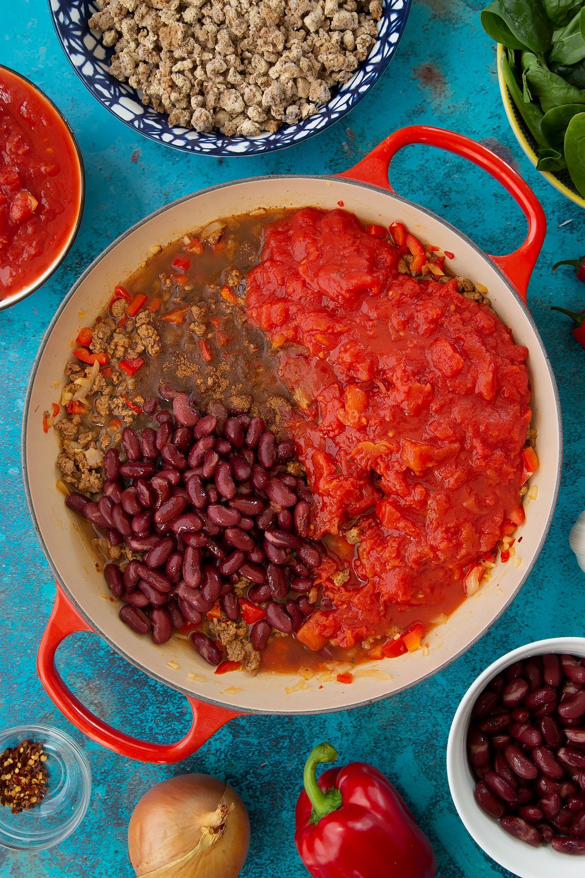 Fried onion, red pepper and vegetarian mince in a pan with kidney beans, tinned tomatoes, cinnamon and stock. Ingredients to make vegetarian mince chilli surround the pan.