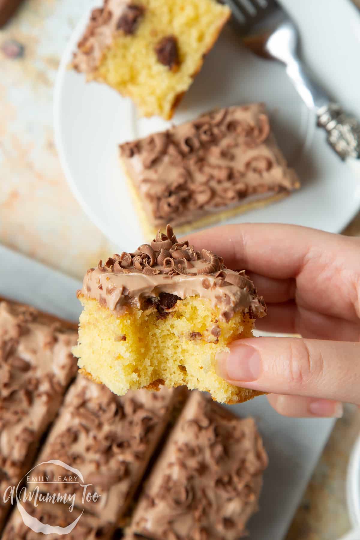 Hand holding a square of chocolate chip tray bake with a bite out of it. More cake on a marble board in the background.