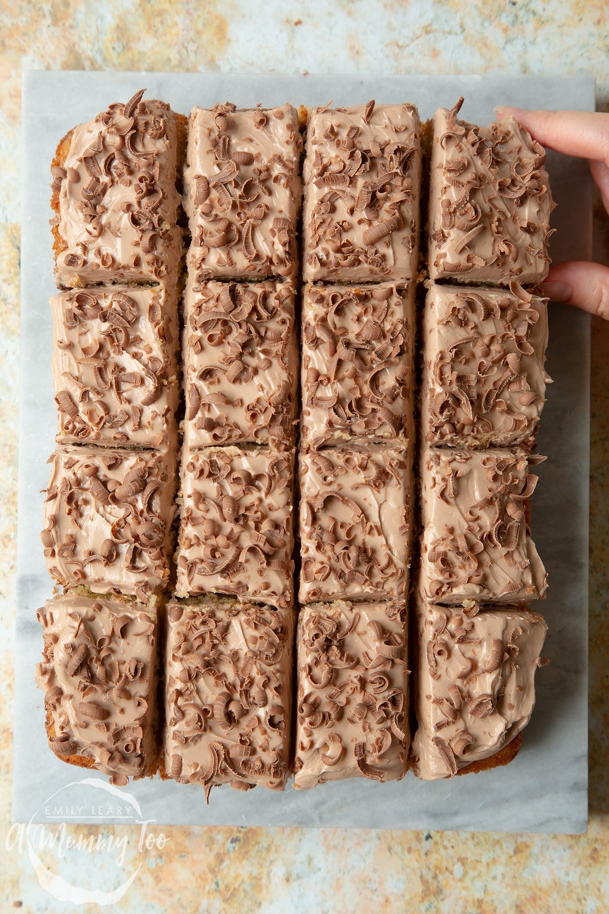 Chocolate chip tray bake on a marble board. A hand reaches to take a square.