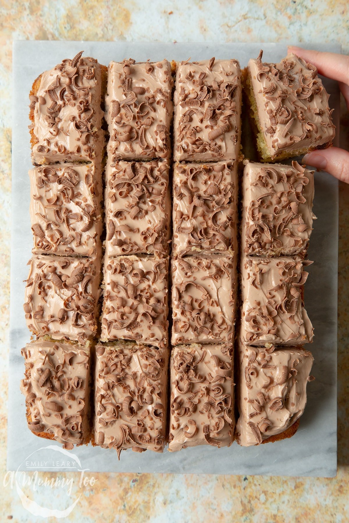 Chocolate chip tray bake on a grey marble board. Fingers reach is to take a piece.