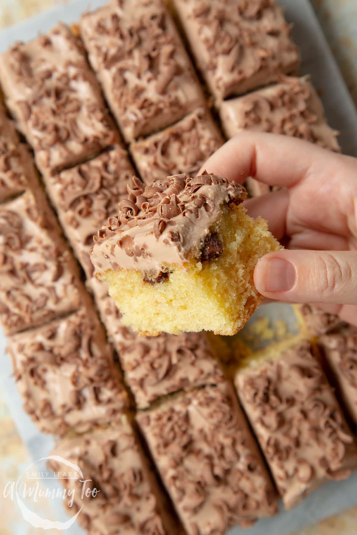 Hand holding a square of chocolate chip tray bake. More cake on a marble board in the background.