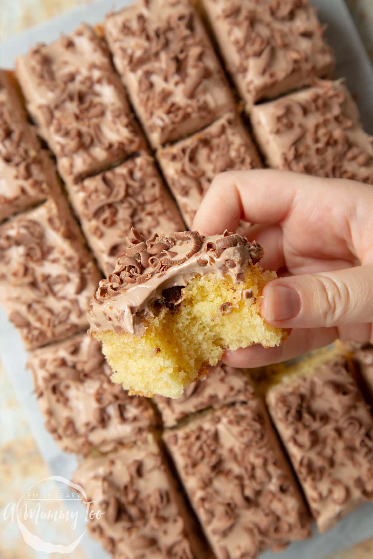 Hand holding a square of chocolate chip tray bake with a bite out of it. More tray bake on a marble board in the background.