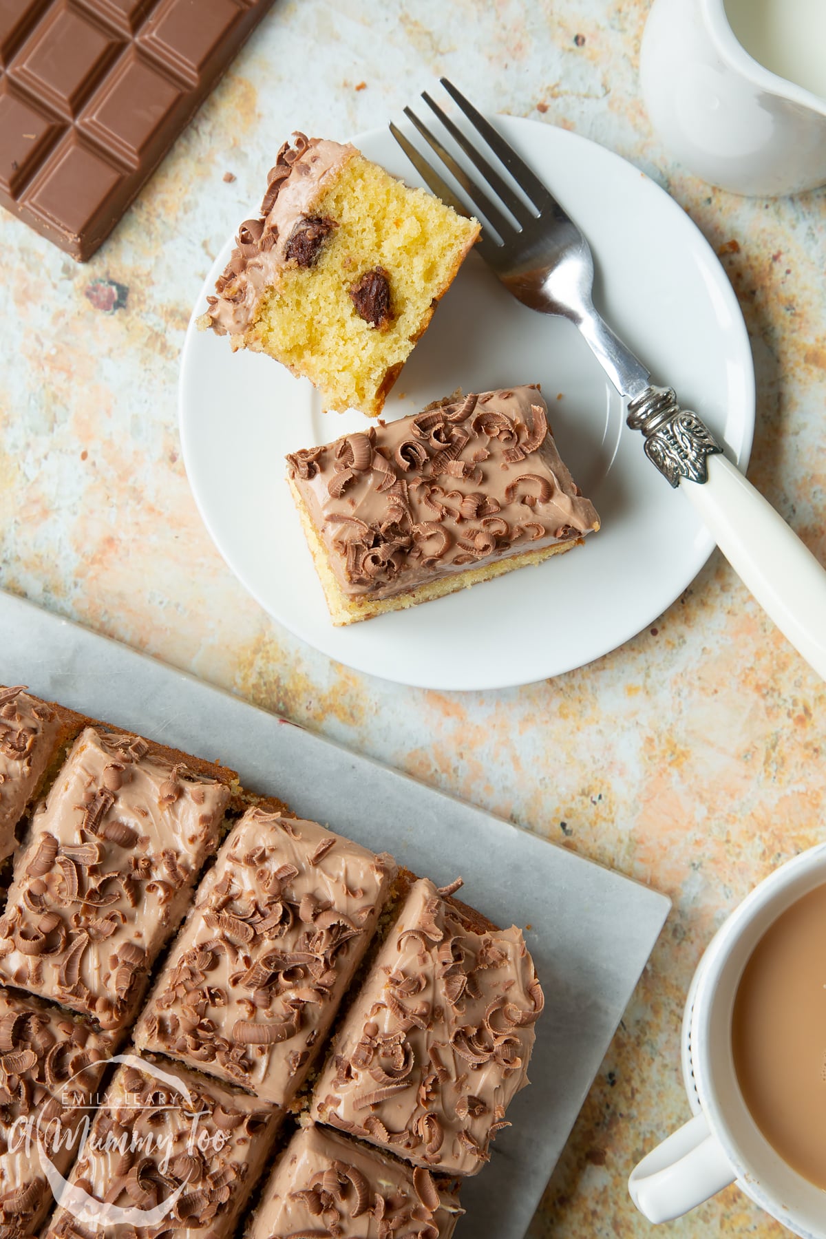 Two squares of chocolate chip tray bake on a white plate with a fork. More is shown on a marble board.