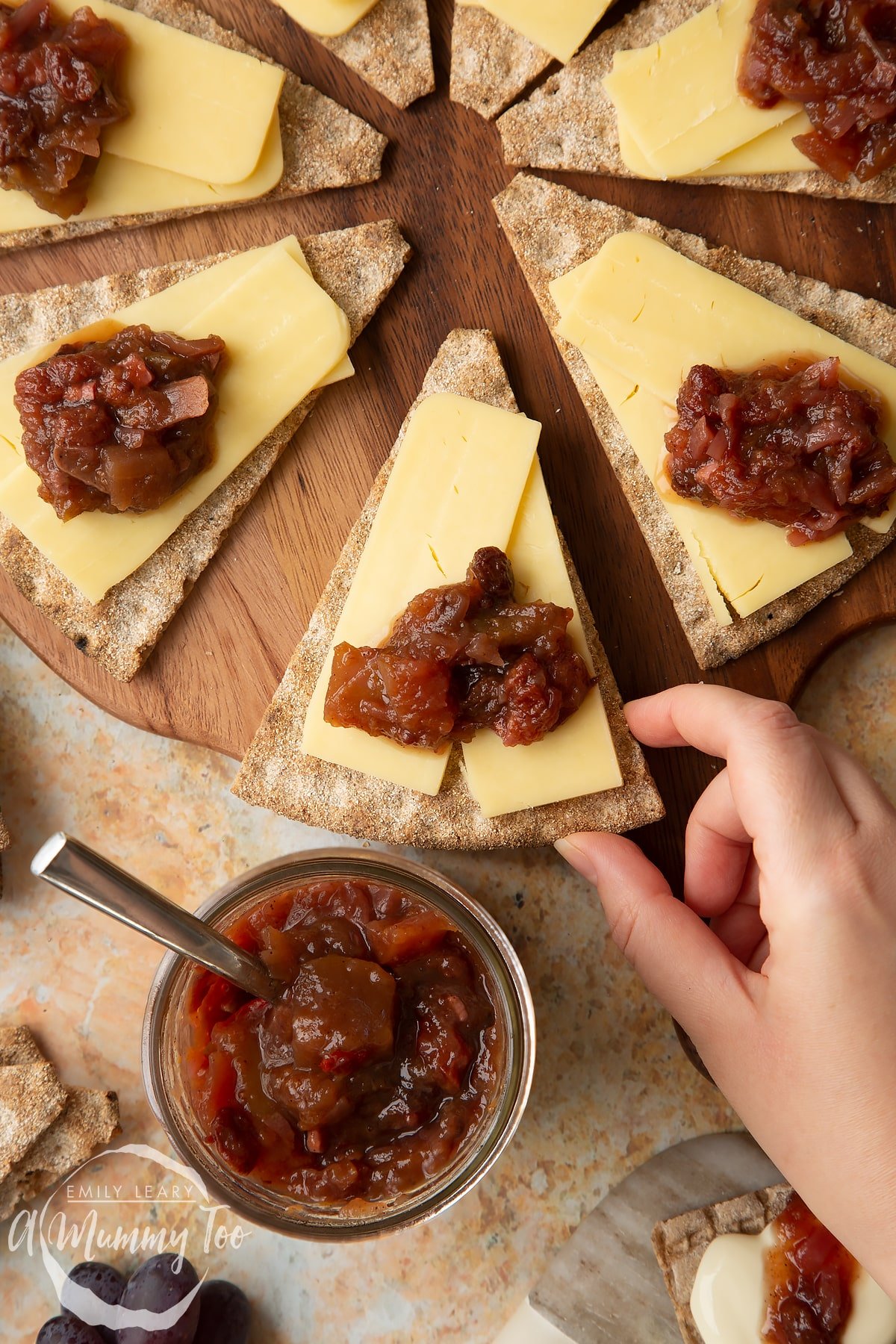 Triangular crackers topped with cheese slices and a fruit chutney recipe on a wooden board, next to a jar of chutney. A hand reaches to take a cracker.