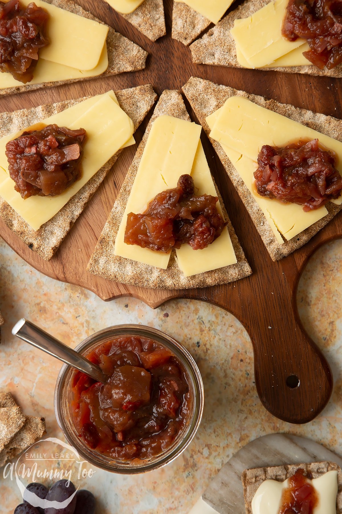 Triangular crackers topped with cheese slices and a fruit chutney recipe on a wooden board, next to a jar of chutney. 