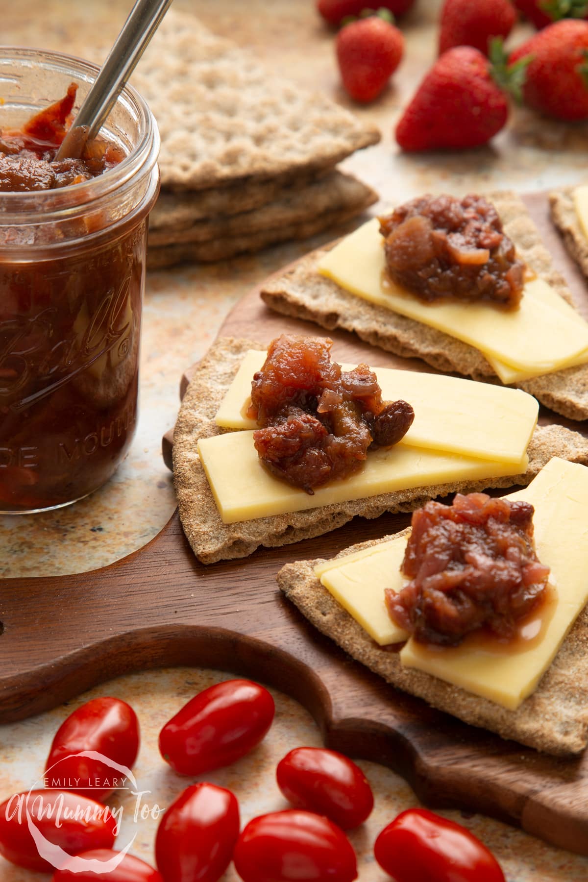 Triangular crackers topped with cheese slices and a fruit chutney recipe on a wooden board, next to a jar of chutney. Salad surrounds the board.