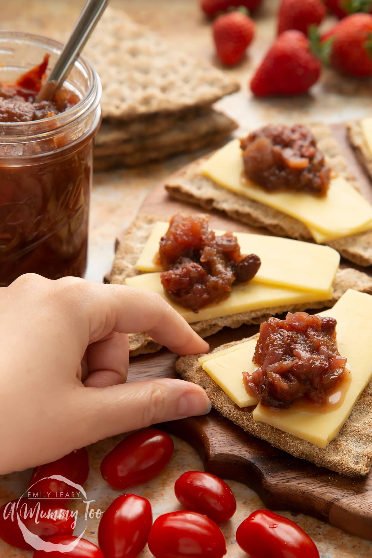 Triangular crackers topped with cheese slices and a fruit chutney recipe on a wooden board, next to a jar of chutney. Salad surrounds the board. A hand reaches to take a cracker.