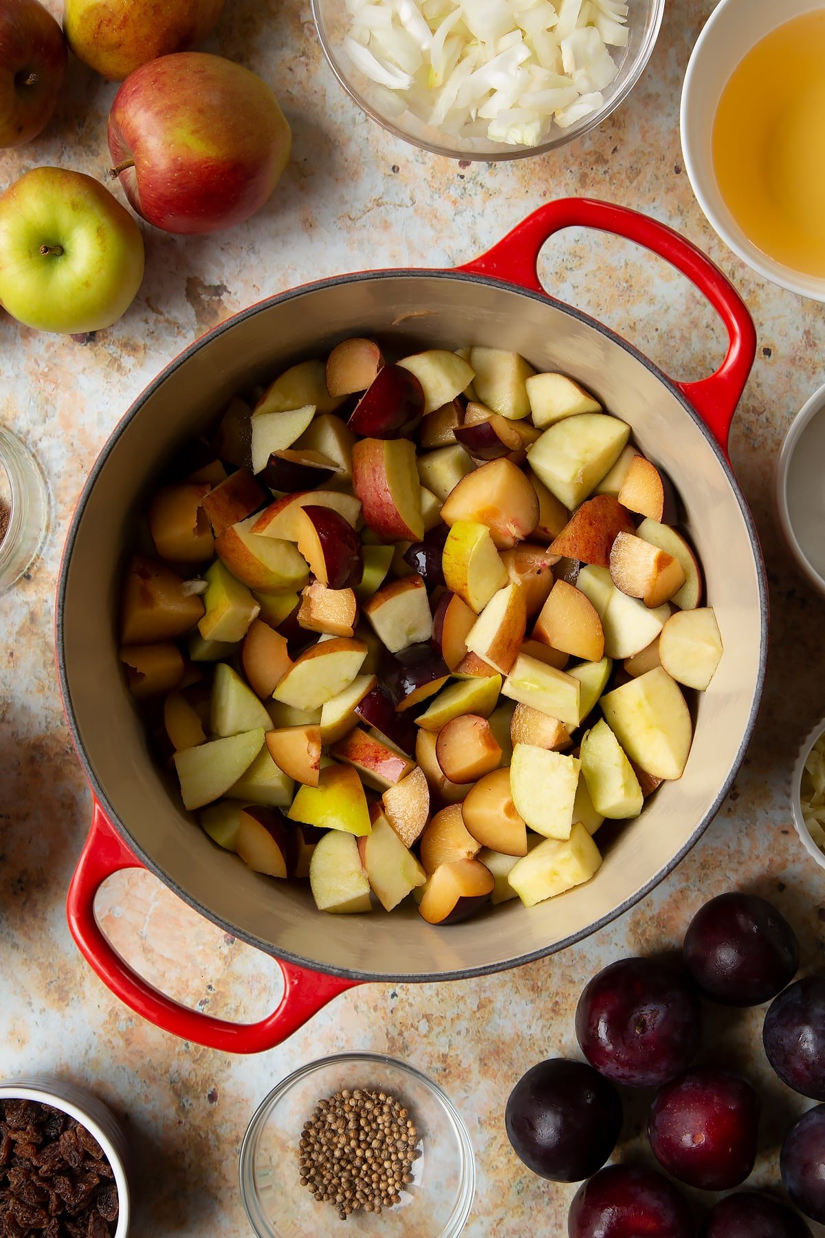 Chopped plums and apples in a large pan. Ingredients to make a fruit chutney recipe surround the pan.