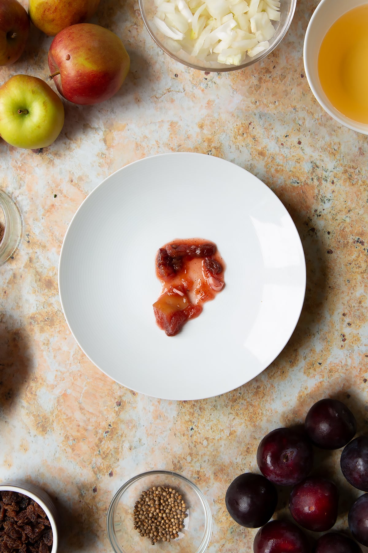 A small amount of fruit chutney on a white plate. Ingredients to make a fruit chutney recipe surround the plate.