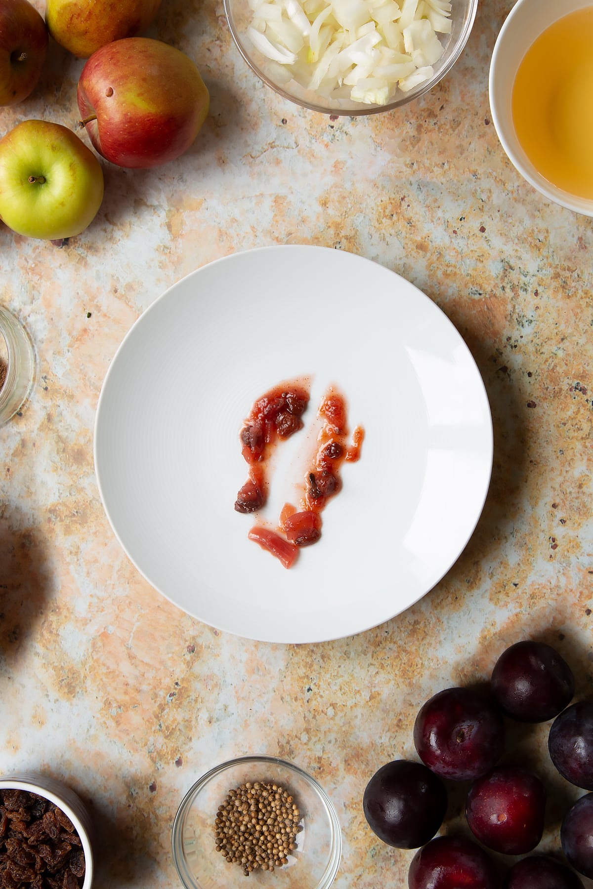 A small amount of fruit chutney on a white plate with a spoon drawn through it. Ingredients to make a fruit chutney recipe surround the plate.