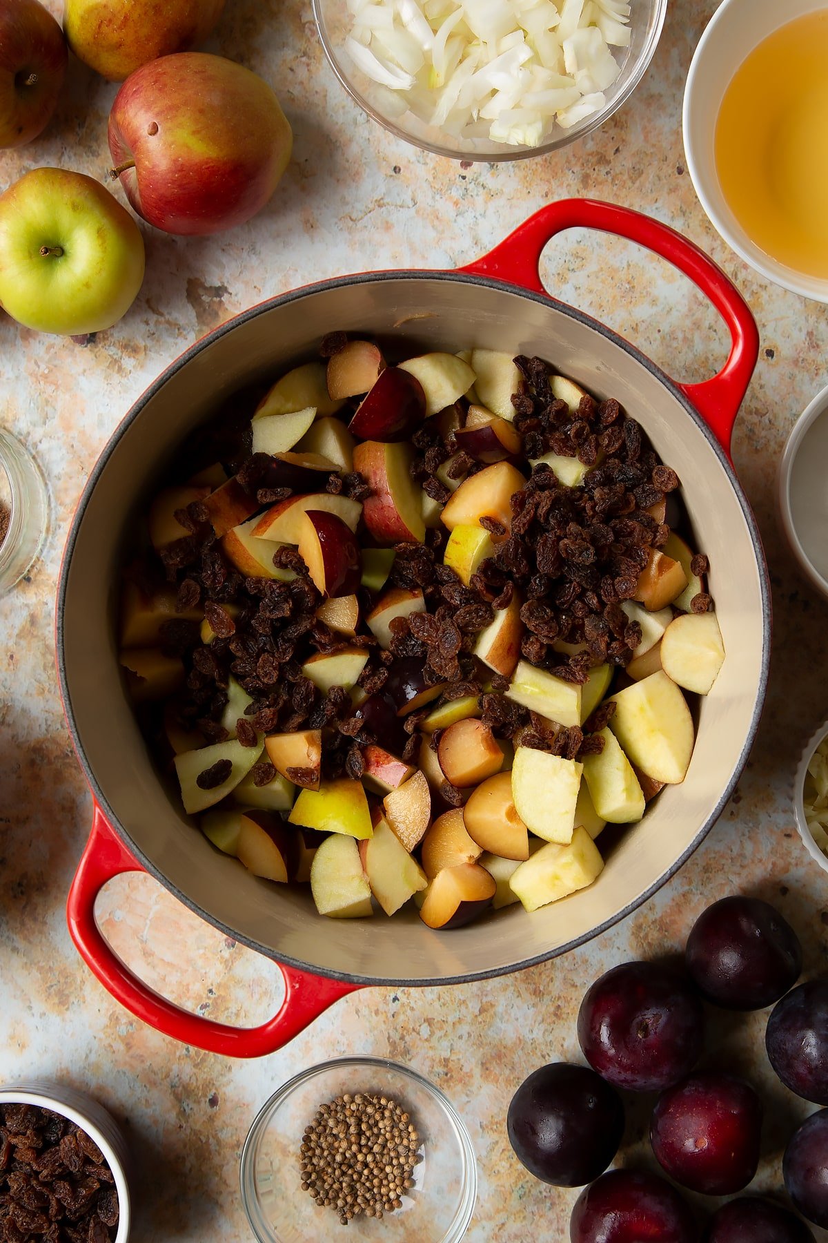 Chopped plums, apples and raisins in a large pan. Ingredients to make a fruit chutney recipe surround the pan.