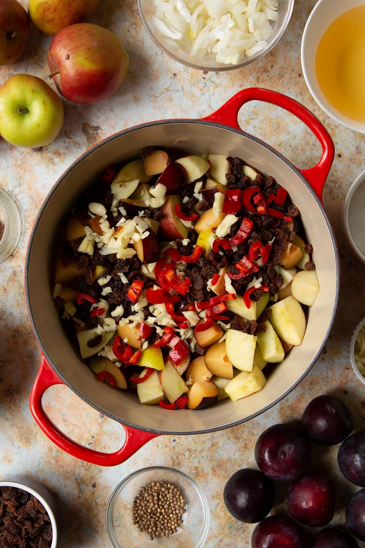 Chopped plums, apples, raisins, chilli and garlic in a large pan. Ingredients to make a fruit chutney recipe surround the pan.