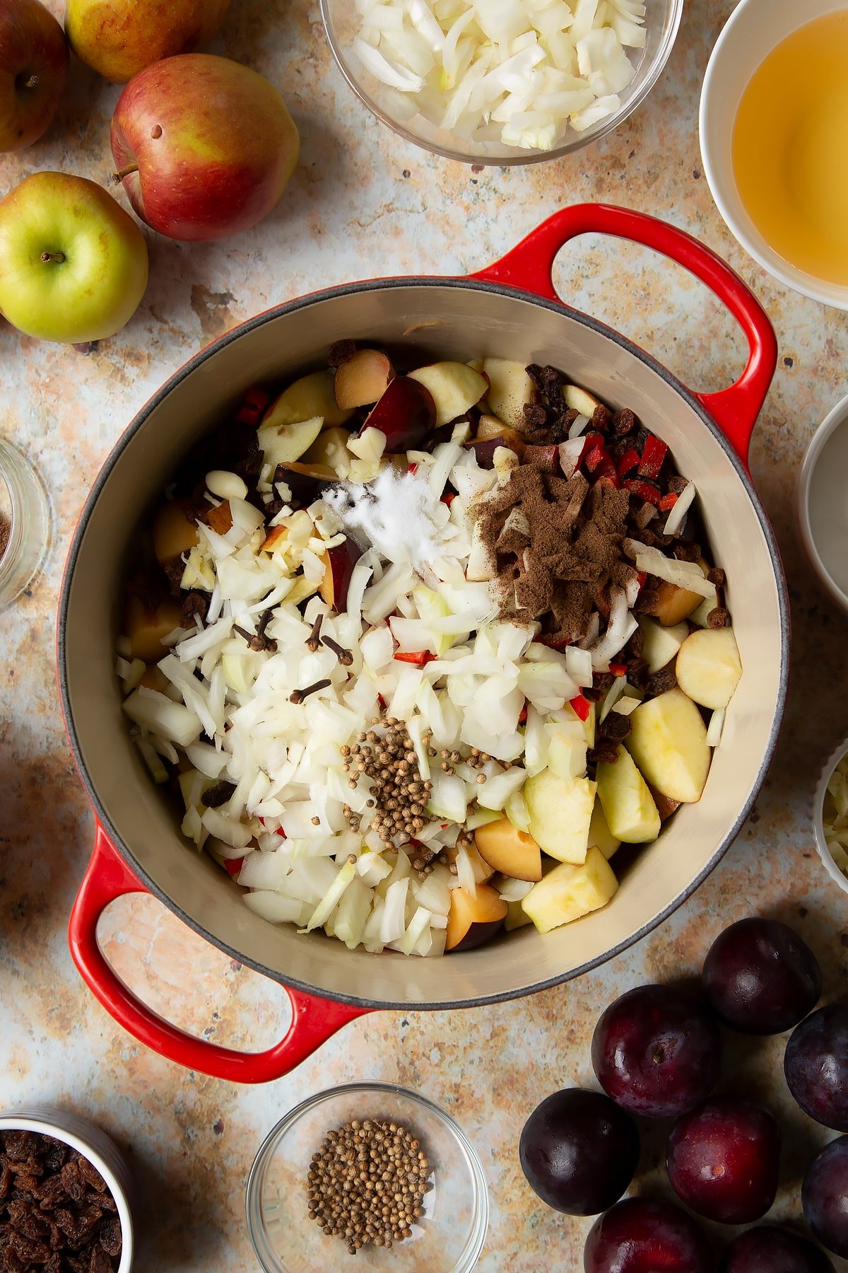 Chopped plums, apples, raisins, chilli, onion and garlic in a large pan. Ingredients to make a fruit chutney recipe surround the pan.