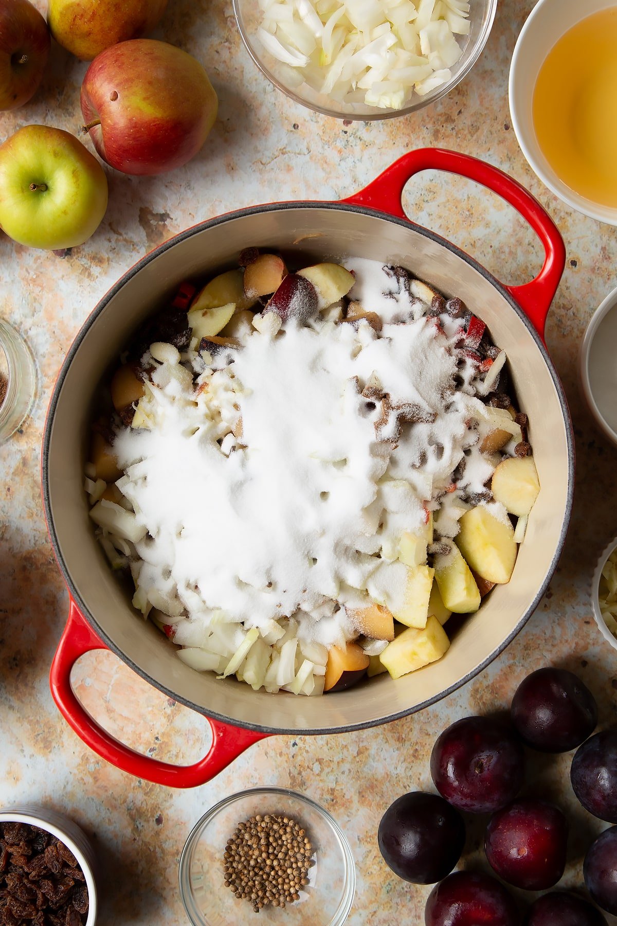 Chopped plums, apples, raisins, chilli, onion, garlic and sugar in a large pan. Ingredients to make a fruit chutney recipe surround the pan.