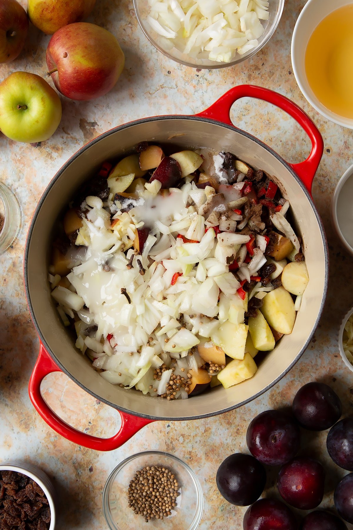 Chopped plums, apples, raisins, chilli, onion, garlic, sugar, vinegar and water in a large pan. Ingredients to make a fruit chutney recipe surround the pan.