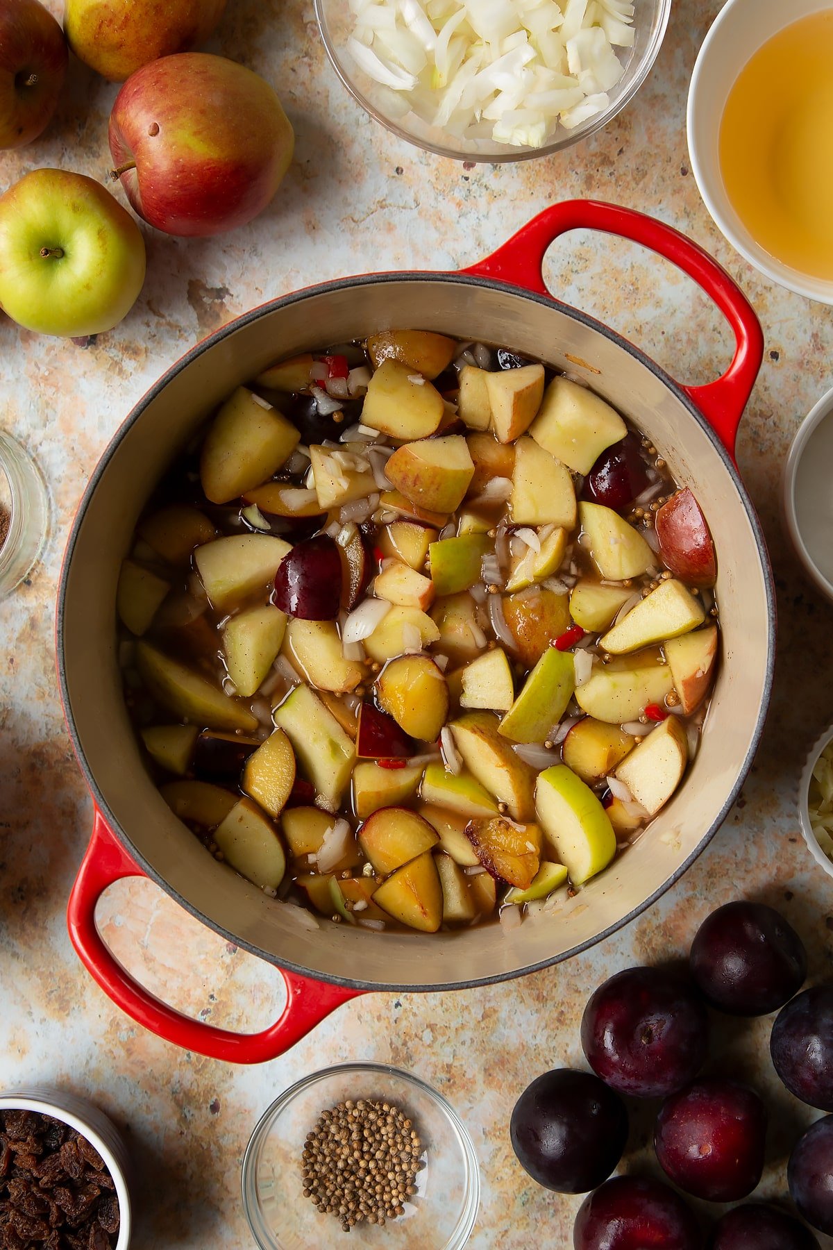 Hot fruit, spices, vinegar and water in a large pan. Ingredients to make a fruit chutney recipe surround the pan.