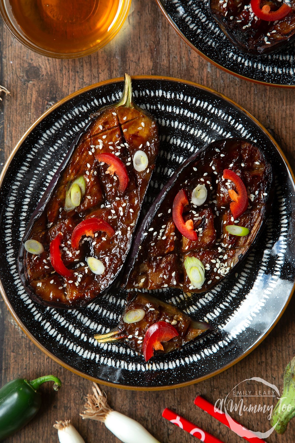 Two slices of miso aubergine on a plate, topped with chilli, springs onions and sesame seeds. One piece has been sliced.