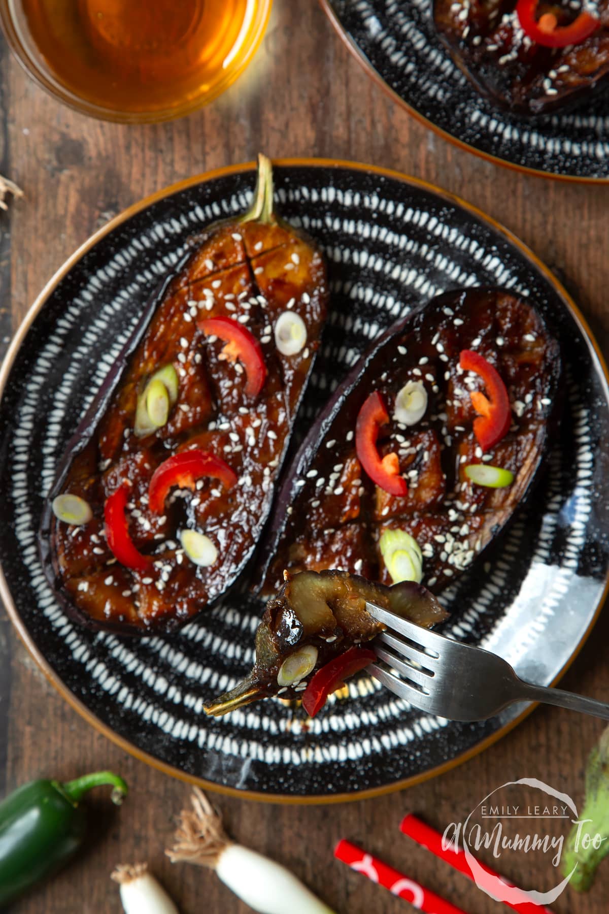 Two slices of miso aubergine on a plate, topped with chilli, springs onions and sesame seeds. A fork holds a piece.