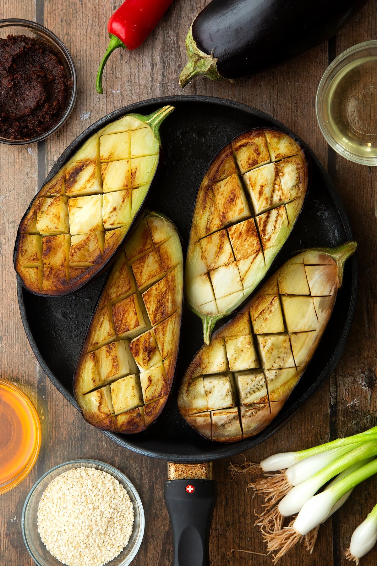 Scored and fried aubergine halves in a pan. Ingredients to make miso aubergine surround the pan.