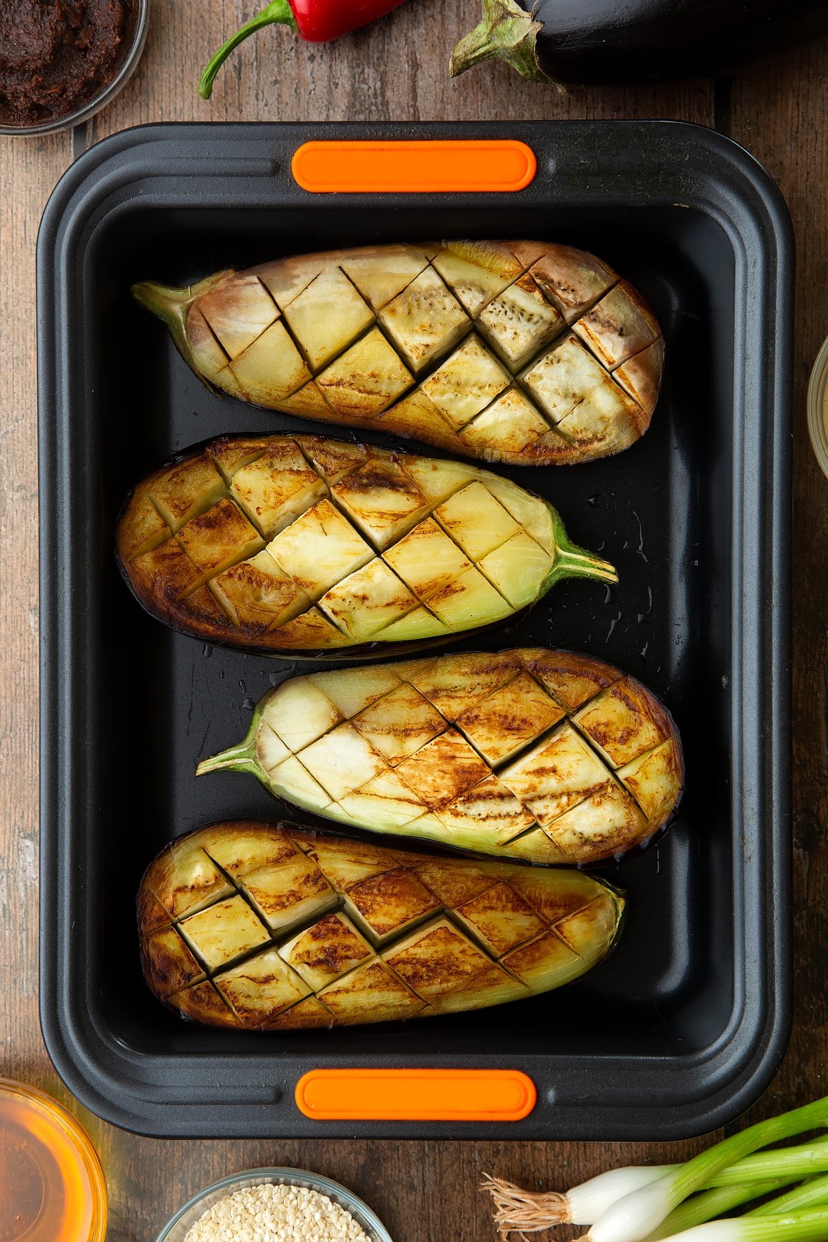 Scored and fried aubergine halves in a tray. Ingredients to make miso aubergine surround the tray.