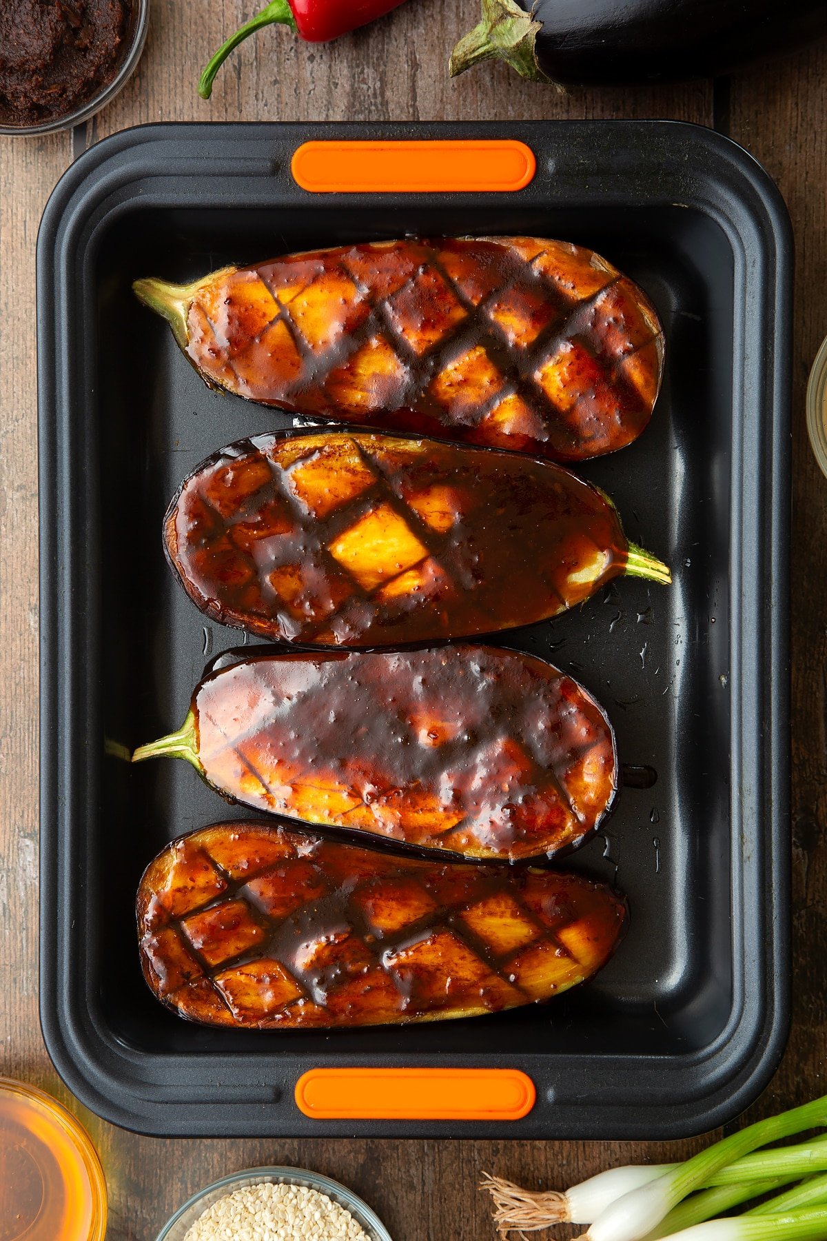 Scored and fried aubergine halves in a tray and covered with a miso glaze. Ingredients to make miso aubergine surround the tray.