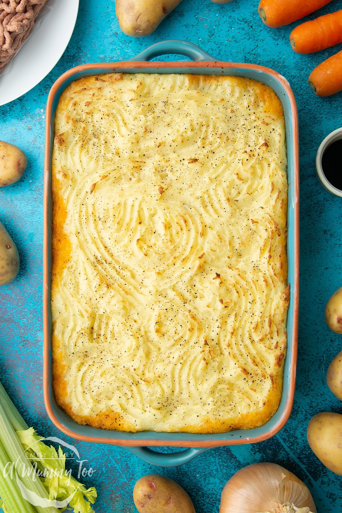 Freshly baked meat free cottage pie in a green roasting dish. Ingredients to make vegan cottage pie surround the dish.